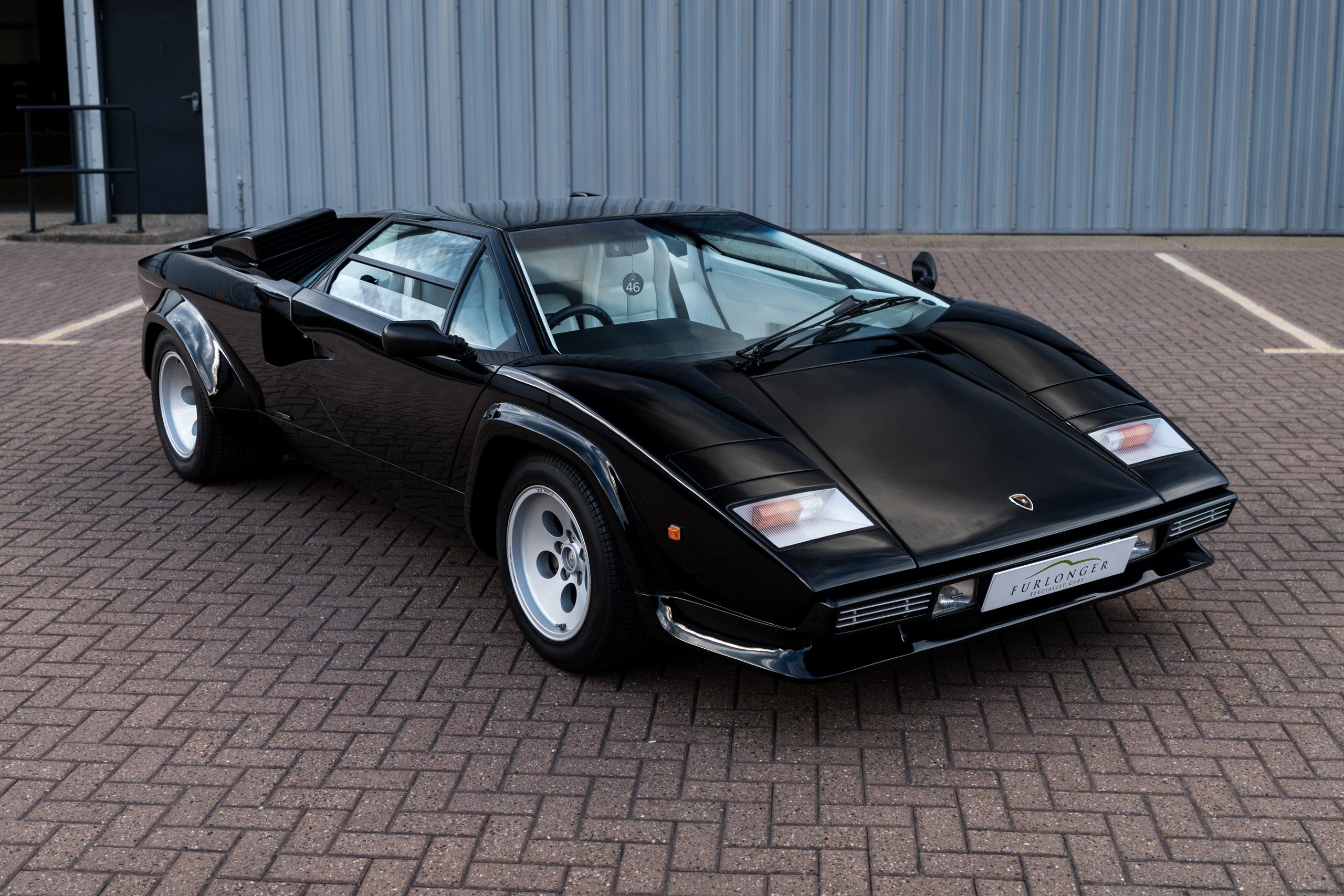Black Lamborghini Countach 5000 S from the front three-quarter angle, with white five-hole wheels, pop-up headlights and a white leather interior visible through the windscreen.
