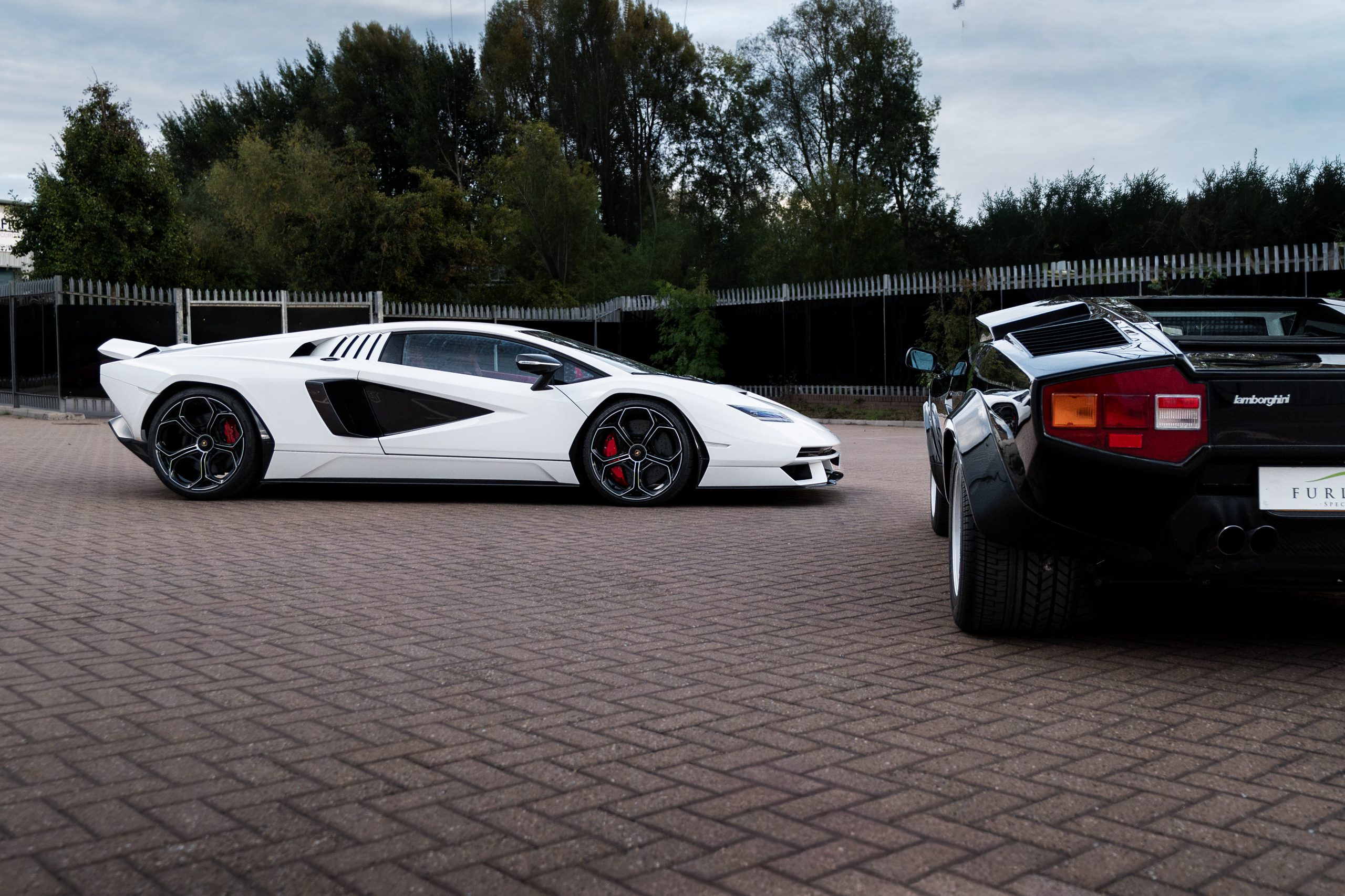 White Countach LPI 800-4 in side profile showing its side air intakes, with the rear of the black Countach 5000 S and its red and amber tail lights in the foreground.